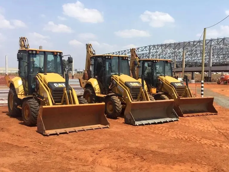 Retroescavadeiras Caterpillar enfileiradas em canteiro de obra, com estrutura metálica ao fundo